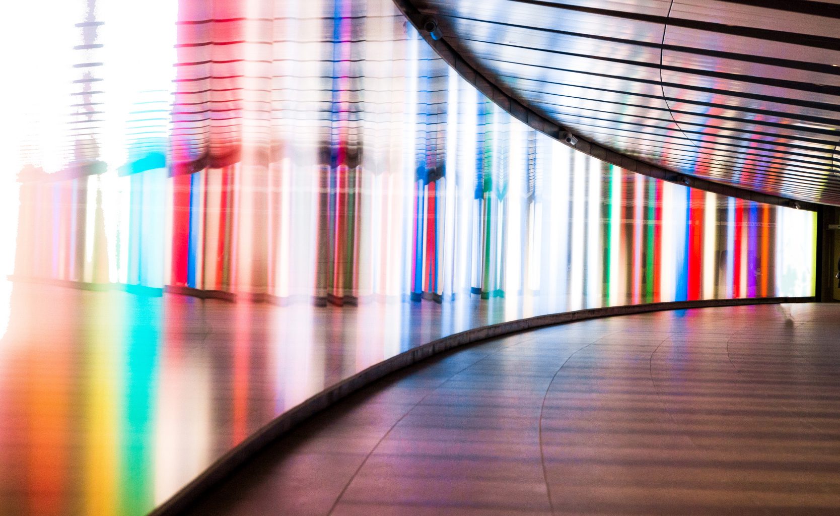 Wide angle color image depicting the front view of a young woman walking through a futuristic subway tunnel in central London, UK. The tunnel is illuminated with multi colored lights. The woman is in the far distance, and the focus is on the cool science fiction architecture of the subway tunnel that curves round into the distance. Room for copy space.
